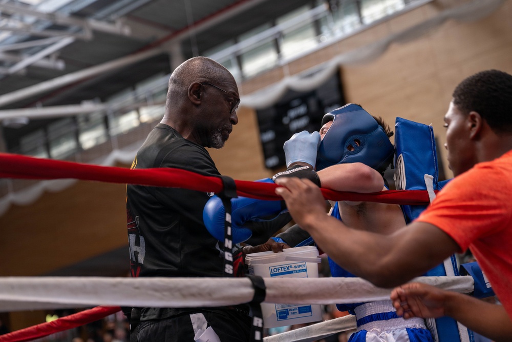 A boxing corner scene showing an older trainer attending to a boxer wearing blue headgear and blue gloves between rounds. The boxer is leaning against the ring ropes while the trainer provides instruction or medical attention. A second cornerman in an orange shirt assists from the right side of the ring.