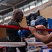 A boxing corner scene showing an older trainer attending to a boxer wearing blue headgear and blue gloves between rounds. The boxer is leaning against the ring ropes while the trainer provides instruction or medical attention. A second cornerman in an orange shirt assists from the right side of the ring.