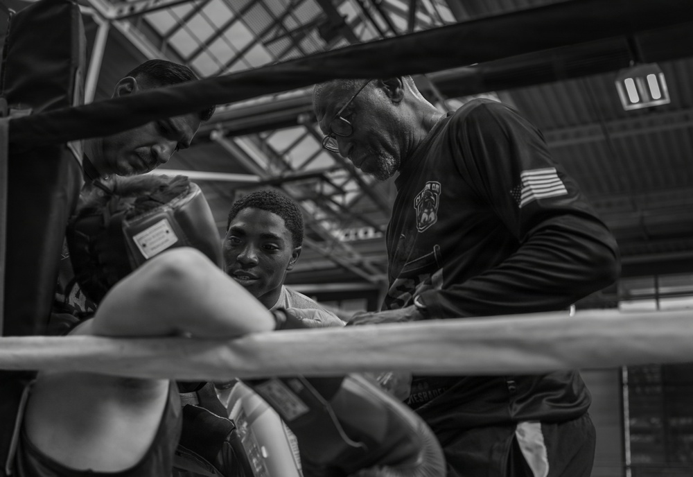 A black-and-white photograph taken from ringside showing a young boxer seated in the corner of a boxing ring during what appears to be a between-rounds break. Two corner men or trainers lean in close, appearing to provide instruction or attend to the fighter. The image is shot through the ropes, giving an intimate, documentary-style perspective.