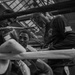 A black-and-white photograph taken from ringside showing a young boxer seated in the corner of a boxing ring during what appears to be a between-rounds break. Two corner men or trainers lean in close, appearing to provide instruction or attend to the fighter. The image is shot through the ropes, giving an intimate, documentary-style perspective.
