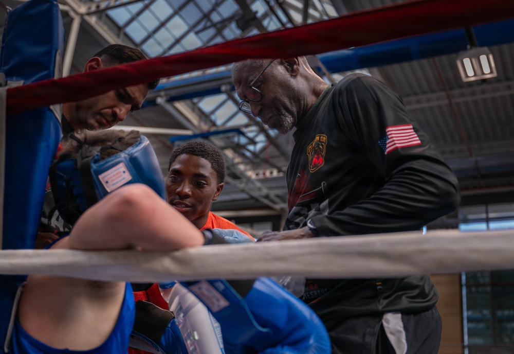 A boxing training or corner session is taking place inside a gymnasium with a glass ceiling. A young boxer in blue and orange gear is receiving instruction or guidance from two corner men between rounds, leaning against the ring ropes. An older man wearing a black long-sleeve shirt with an American flag patch appears to be the head trainer or coach.