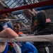 A boxing training or corner session is taking place inside a gymnasium with a glass ceiling. A young boxer in blue and orange gear is receiving instruction or guidance from two corner men between rounds, leaning against the ring ropes. An older man wearing a black long-sleeve shirt with an American flag patch appears to be the head trainer or coach.