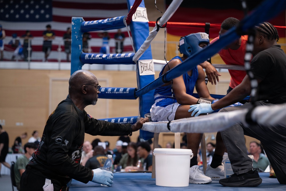 A boxing match corner scene showing a boxer in blue gear resting on a stool between rounds while corner men attend to them. Two cornermen wearing blue latex gloves are providing care and instruction to the boxer inside a Ringside-branded boxing ring. An American flag is visible in the background of what appears to be an indoor arena with spectators.