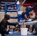 A boxing match corner scene showing a boxer in blue gear resting on a stool between rounds while corner men attend to them. Two cornermen wearing blue latex gloves are providing care and instruction to the boxer inside a Ringside-branded boxing ring. An American flag is visible in the background of what appears to be an indoor arena with spectators.