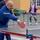1ID Band Energizes Bolesławiec A bald middle-aged man wearing a blue and gold tracksuit is playing table tennis outdoors, captured mid-stroke with a red paddle and white ball visible. The event appears to be a public or community outdoor table tennis activity, with a second table and a young girl playing in the background. The table is branded 'POLSPORT BIELSKO-BIAŁA', suggesting a Polish sporting event or community gathering.