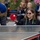 1ID Band Energizes Bolesławiec A young woman using a wheelchair is playing table tennis outdoors, holding a red paddle with a white ball visible near it. She is wearing sunglasses, a black jacket with a green lanyard, and appears focused and engaged in the game. Several spectators are seated on benches in the background watching the activity.