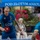 1ID Band Energizes Bolesławiec Two men are playing table tennis outdoors in what appears to be a restaurant or café courtyard called 'Pod Złotym Aniołem' (Under the Golden Angel), likely in Poland. The foreground player wears a blue 'LOB Team' jersey and holds a red paddle mid-play. An elderly woman watches from a bench in the background surrounded by colorful flowers.
