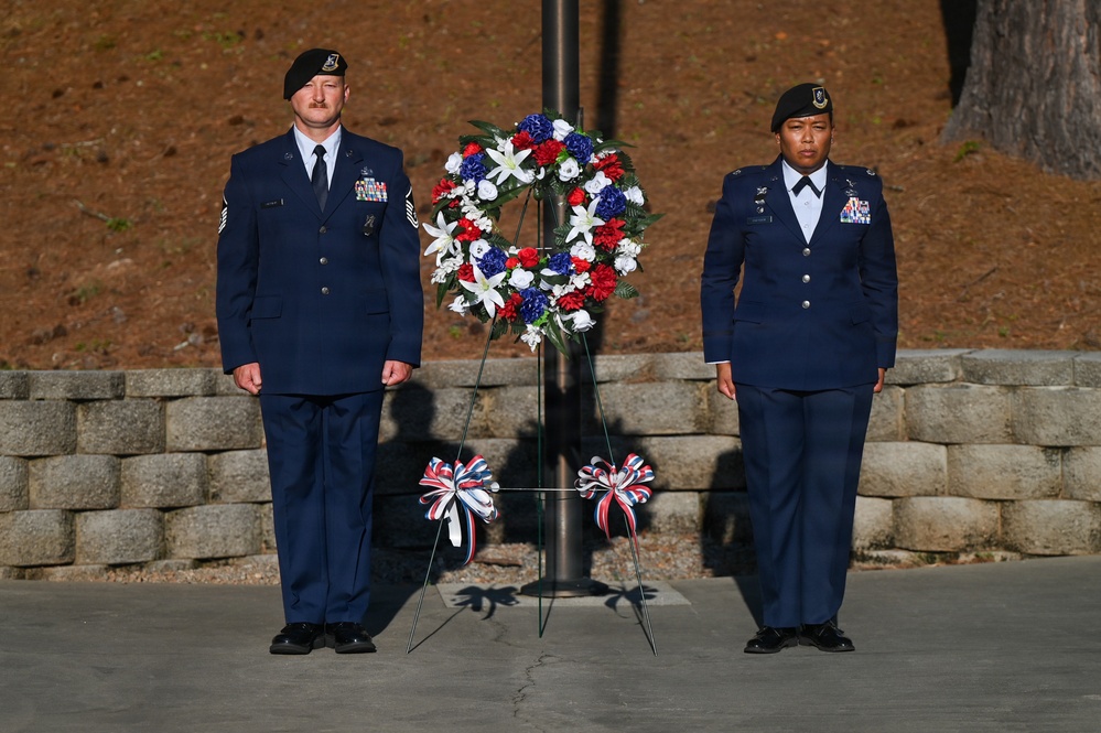 Two U.S. Air Force personnel in dress blue uniforms stand at attention flanking a red, white, and blue memorial wreath mounted on a stand. The scene appears to be a formal military memorial or wreath-laying ceremony, likely at an outdoor memorial site. Both individuals wear green berets and display service ribbons and badges on their uniforms.
