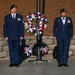 Two U.S. Air Force personnel in dress blue uniforms stand at attention flanking a red, white, and blue memorial wreath mounted on a stand. The scene appears to be a formal military memorial or wreath-laying ceremony, likely at an outdoor memorial site. Both individuals wear green berets and display service ribbons and badges on their uniforms.