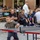 1ID Band Energizes Bolesławiec A young boy wearing glasses and a Hugo-branded hoodie is playing table tennis outdoors in what appears to be a public square or plaza. Behind him, several adults and a teenage girl are seated watching, with an outdoor seating area for a venue called 'The Pub' visible in the background. The scene suggests a casual public recreational event or setup in a European town center.