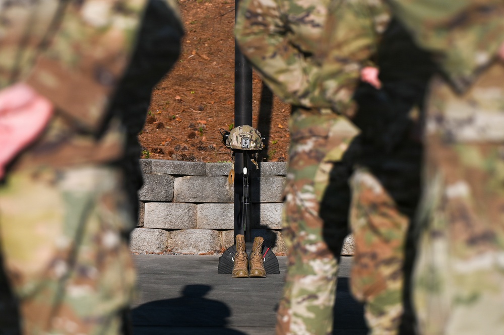 The image depicts a military memorial display, commonly known as a 'Fallen Soldier Battle Cross' or 'Battle Cross Memorial,' consisting of boots, a rifle, and a helmet arranged on a stand. Soldiers in camouflage uniforms are visible in the foreground, blurred, appearing to stand in formation around the memorial. This type of display is traditionally used during memorial ceremonies to honor fallen service members.