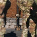 The image depicts a military memorial display, commonly known as a 'Fallen Soldier Battle Cross' or 'Battle Cross Memorial,' consisting of boots, a rifle, and a helmet arranged on a stand. Soldiers in camouflage uniforms are visible in the foreground, blurred, appearing to stand in formation around the memorial. This type of display is traditionally used during memorial ceremonies to honor fallen service members.