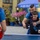 1ID Band Energizes Bolesławiec An adult man is coaching a young boy in table tennis outdoors, guiding his paddle stroke from behind. The scene takes place in what appears to be a town square or plaza, with a blue umbrella canopy visible in the background bearing partial text 'POD ZŁO... AN...'. A third player holding a red paddle is visible in the foreground, facing them across the table.