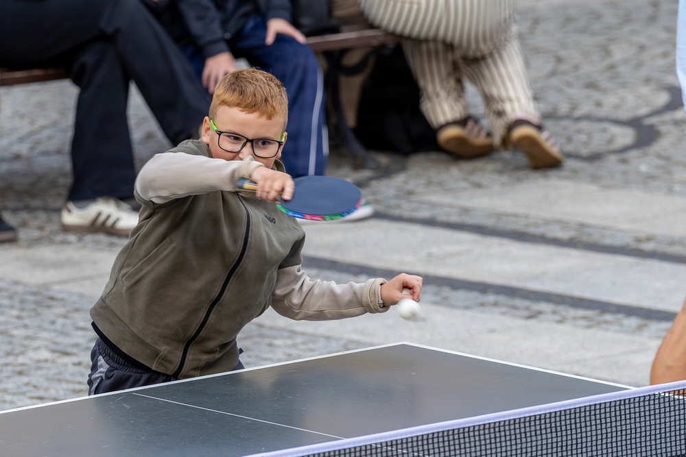 A young red-haired boy wearing glasses and a gray-green jacket is actively playing table tennis outdoors. He is mid-swing, making contact with a white ping-pong ball using a colorful blue and pink paddle. Several adults are seated on a bench in the blurred background, watching the activity.