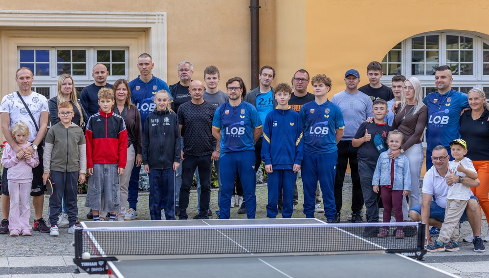 A group of approximately 25-30 people, including adults and children, pose for a group photo outdoors in front of a yellow building. Several individuals are wearing blue 'LOB Team' branded athletic uniforms, suggesting this is a sports team or club gathering. A ping-pong (table tennis) table with a Tibhar-branded net is prominently visible in the foreground.