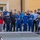 1ID Band Energizes Bolesławiec A group of approximately 25-30 people, including adults and children, pose for a group photo outdoors in front of a yellow building. Several individuals are wearing blue 'LOB Team' branded athletic uniforms, suggesting this is a sports team or club gathering. A ping-pong (table tennis) table with a Tibhar-branded net is prominently visible in the foreground.