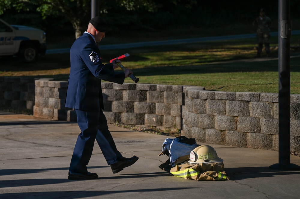 A U.S. Air Force Master Sergeant in dress blues uniform is walking while carrying what appears to be a fire hose nozzle or similar equipment. On the ground nearby lies a set of firefighter turnout gear including a helmet and jacket, suggesting a ceremonial or memorial event. The setting appears to be an outdoor paved area on a military installation.
