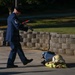 A U.S. Air Force Master Sergeant in dress blues uniform is walking while carrying what appears to be a fire hose nozzle or similar equipment. On the ground nearby lies a set of firefighter turnout gear including a helmet and jacket, suggesting a ceremonial or memorial event. The setting appears to be an outdoor paved area on a military installation.