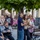 1ID Band Energizes Bolesławiec A group of people gathered in an outdoor public square or plaza, likely watching a performance or event out of frame. The scene takes place beneath a large leafy tree, with colorful flower beds and storefronts visible in the background. The atmosphere appears casual and communal, suggesting a public event or street performance.
