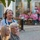 1ID Band Energizes Bolesławiec Three women are gathered in an outdoor public square or pedestrian area, likely watching an event or performance off-camera. The scene appears to be a warm-weather afternoon in what looks like a European town center, with pink flowers, café seating, and storefronts visible in the background. The women display varying expressions of attentiveness and mild interest.