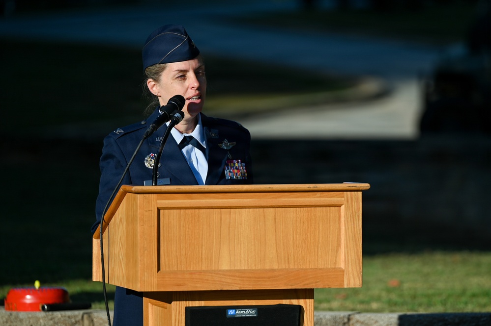 A female U.S. Air Force officer in dress blues is delivering a speech at an outdoor podium. She is wearing a service cap, multiple medals and ribbons, and pilot wings, indicating a senior rank. The setting appears to be a formal military ceremony held outdoors during daylight hours.