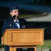 A female U.S. Air Force officer in dress blues is delivering a speech at an outdoor podium. She is wearing a service cap, multiple medals and ribbons, and pilot wings, indicating a senior rank. The setting appears to be a formal military ceremony held outdoors during daylight hours.