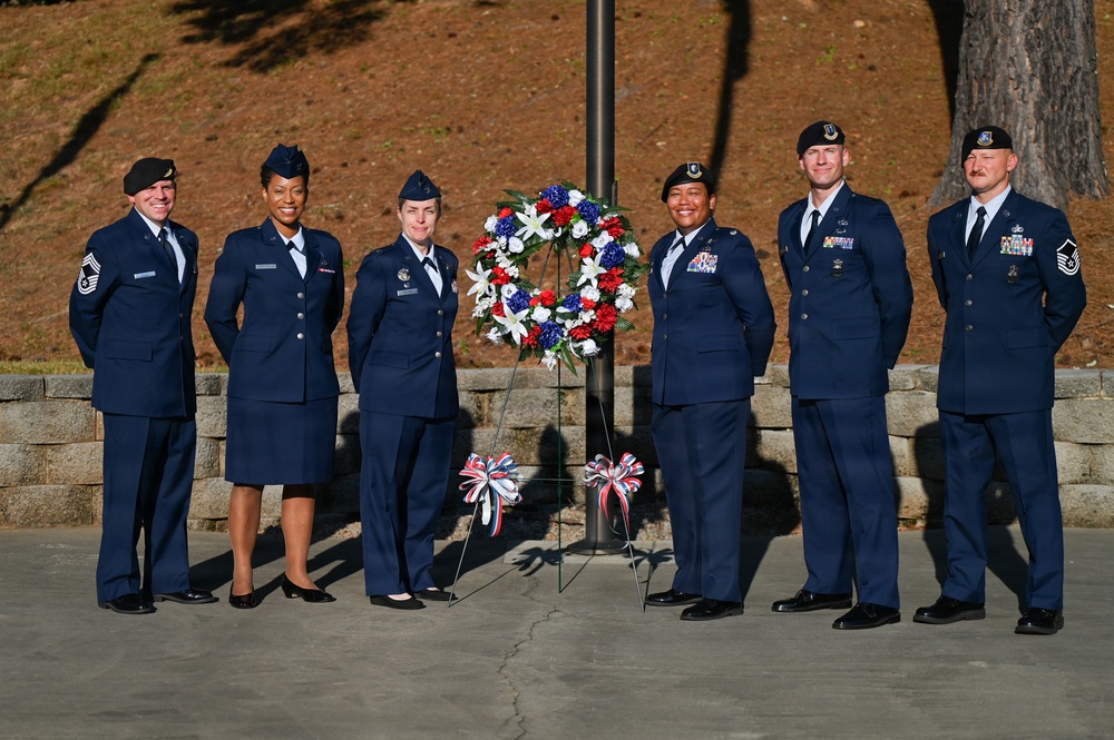 Six United States Air Force personnel in dress blue uniforms are posed together at what appears to be a memorial or ceremonial event. They are standing in front of a patriotic wreath featuring red, white, and blue flowers mounted on a stand near a flagpole. The setting appears to be an outdoor memorial area with a stone wall backdrop.