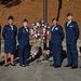 Six United States Air Force personnel in dress blue uniforms are posed together at what appears to be a memorial or ceremonial event. They are standing in front of a patriotic wreath featuring red, white, and blue flowers mounted on a stand near a flagpole. The setting appears to be an outdoor memorial area with a stone wall backdrop.