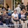 1ID Band Energizes Bolesławiec A group of people are seated on a bench in an outdoor public square, with three adult women and a young girl in the foreground engaged in conversation. Behind them is a busy outdoor seating area belonging to an establishment called 'The Pub', decorated with hop-themed signage in gold and black. The scene appears to be a lively public gathering space, likely in a European city.