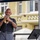 1ID Band Energizes Bolesławiec A bald man is performing outdoors, playing a brass trumpet under a blue canopy tent. He is positioned on what appears to be a small stage with a music stand and microphone visible nearby. The setting appears to be a public outdoor venue with a European-style building visible in the background.