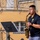 1ID Band Energizes Bolesławiec A male musician is performing outdoors on a stage, playing a brass alto saxophone while reading sheet music from a stand. He is wearing a black polo shirt with a shield logo and the text '1D BAND', suggesting affiliation with a military or organized band. The background features a yellow European-style building with ornate iron railings.