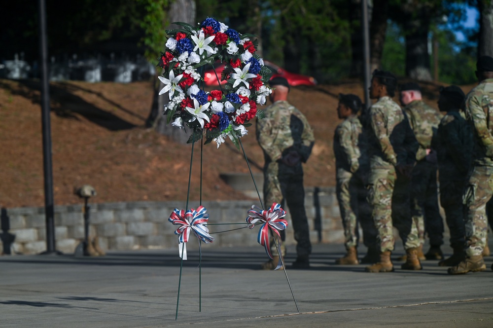 A military memorial ceremony is taking place outdoors, featuring a prominent patriotic wreath with red, white, and blue flowers displayed on a stand in the foreground. Several soldiers in camouflage uniforms (OCP/MultiCam pattern) are standing in formation or at ease in the background. The setting appears to be a cemetery or memorial grounds with stone walls and pine trees visible.