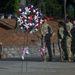 A military memorial ceremony is taking place outdoors, featuring a prominent patriotic wreath with red, white, and blue flowers displayed on a stand in the foreground. Several soldiers in camouflage uniforms (OCP/MultiCam pattern) are standing in formation or at ease in the background. The setting appears to be a cemetery or memorial grounds with stone walls and pine trees visible.