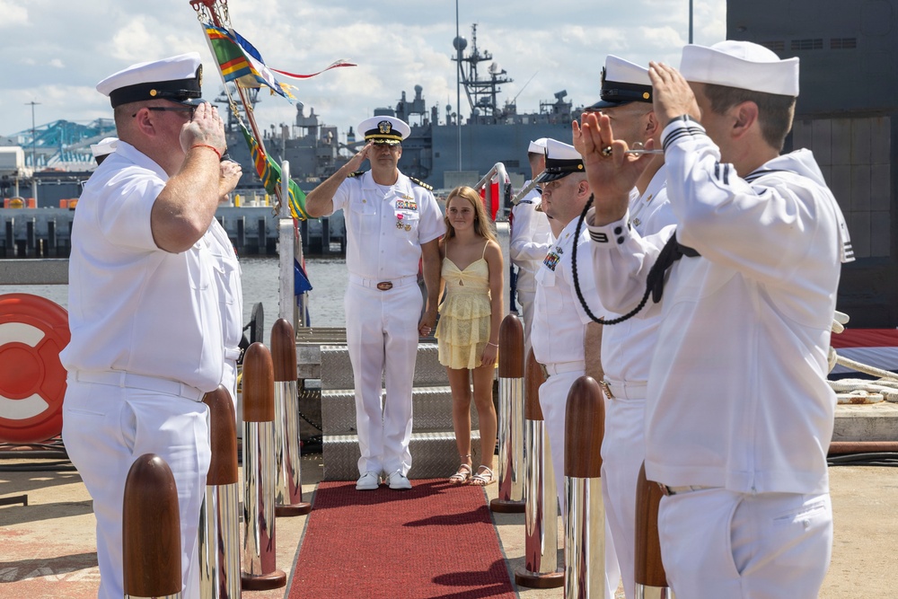 A U.S. Navy ceremonial event taking place on a pier or dock, likely a change of command or arrival/departure ceremony. Multiple sailors in white dress uniforms are forming a sideboy honor guard, rendering salutes along a red carpet. A senior officer wearing medals and a young civilian girl are walking through the ceremonial corridor.
