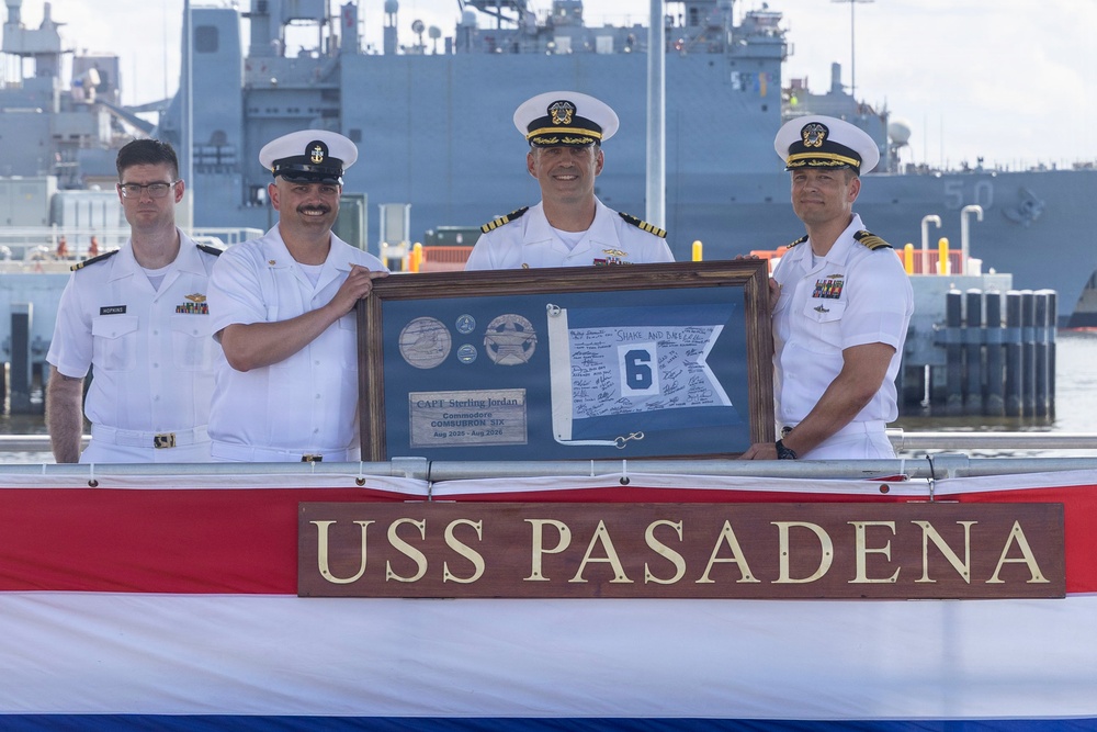 Four U.S. Navy personnel in white dress uniforms are posing aboard or in front of the USS Pasadena, holding a framed shadow box presentation. The framed display appears to be a commemorative gift for CAPT Sterling Jordan, Commodore of COMSUBRON SIX (Submarine Squadron Six), covering the period August 2020 to August 2026. A large gray naval vessel is visible in the background at what appears to be a naval base pier.