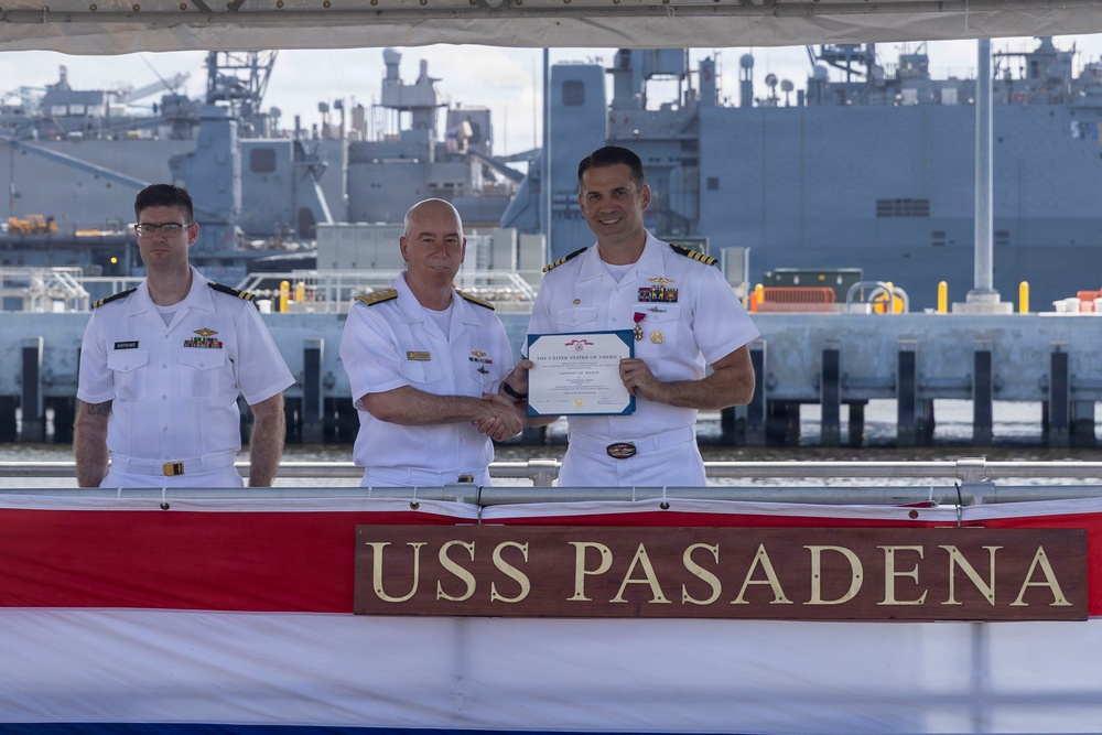 A military ceremony is taking place aboard or in front of the USS Pasadena, a U.S. Navy vessel. Two naval officers in white dress uniforms are shaking hands while one presents a certificate or award to the other, with a third officer standing at attention in the background. The setting appears to be a naval pier or shipyard with large vessels visible in the background.