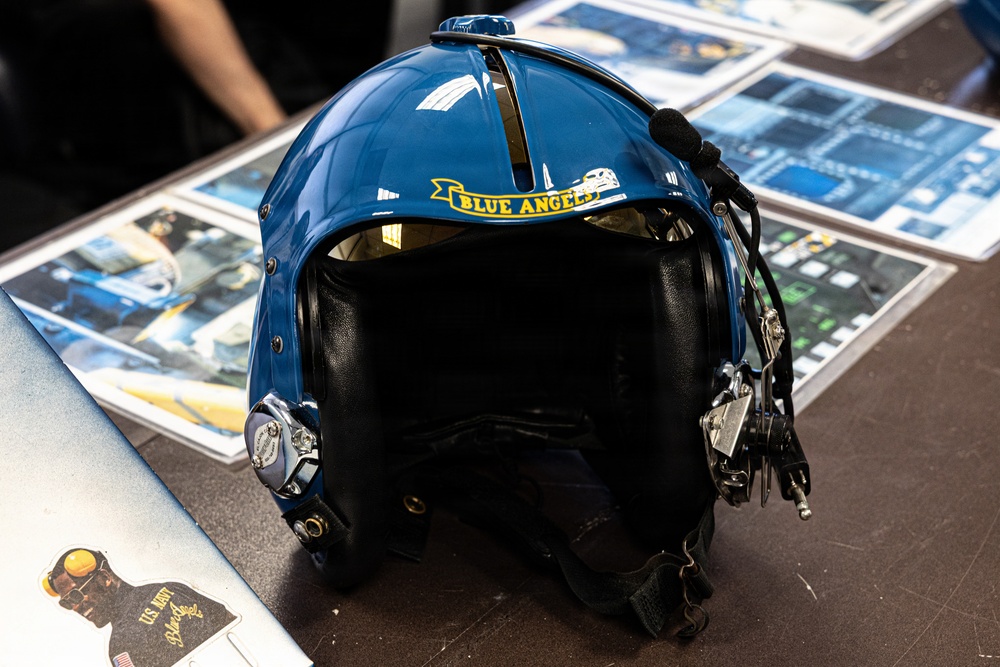 A Blue Angels U.S. Navy flight helmet is displayed on a table, featuring a glossy blue shell with yellow 'Blue Angels' banner logo. The helmet includes communication equipment such as a microphone boom and audio connectors on the right side. It is surrounded by printed photographs and promotional materials related to the Blue Angels flight demonstration squadron.