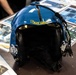 A Blue Angels U.S. Navy flight helmet is displayed on a table, featuring a glossy blue shell with yellow 'Blue Angels' banner logo. The helmet includes communication equipment such as a microphone boom and audio connectors on the right side. It is surrounded by printed photographs and promotional materials related to the Blue Angels flight demonstration squadron.
