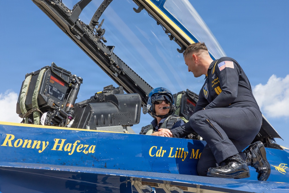 A Blue Angels Navy flight demonstration squadron aircraft is shown on the ground with a pilot seated in the cockpit wearing a blue flight helmet and oxygen mask. A uniformed Navy officer is crouching on the aircraft's fuselage, appearing to brief or speak with the seated pilot. The aircraft's fuselage displays the names 'Ronny Hafeza' and 'Cdr Lilly Mo[rgan or similar]' in gold lettering on the blue paint scheme.