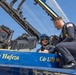 A Blue Angels Navy flight demonstration squadron aircraft is shown on the ground with a pilot seated in the cockpit wearing a blue flight helmet and oxygen mask. A uniformed Navy officer is crouching on the aircraft's fuselage, appearing to brief or speak with the seated pilot. The aircraft's fuselage displays the names 'Ronny Hafeza' and 'Cdr Lilly Mo[rgan or similar]' in gold lettering on the blue paint scheme.