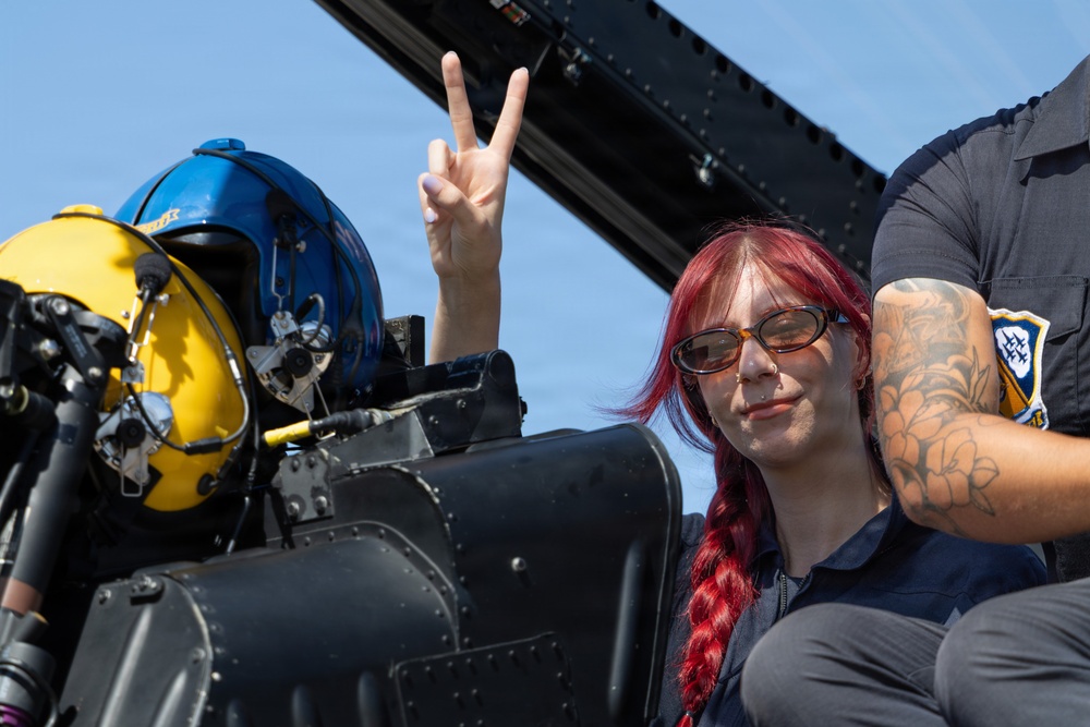 A woman with red braided hair and amber-tinted sunglasses is seated in or near a military aircraft cockpit, making a peace/victory sign with her hand. A blue and yellow flight helmet is visible on the cockpit equipment beside her. A second person with a tattooed arm and a uniform patch is partially visible on the right side of the frame.