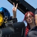A woman with red braided hair and amber-tinted sunglasses is seated in or near a military aircraft cockpit, making a peace/victory sign with her hand. A blue and yellow flight helmet is visible on the cockpit equipment beside her. A second person with a tattooed arm and a uniform patch is partially visible on the right side of the frame.