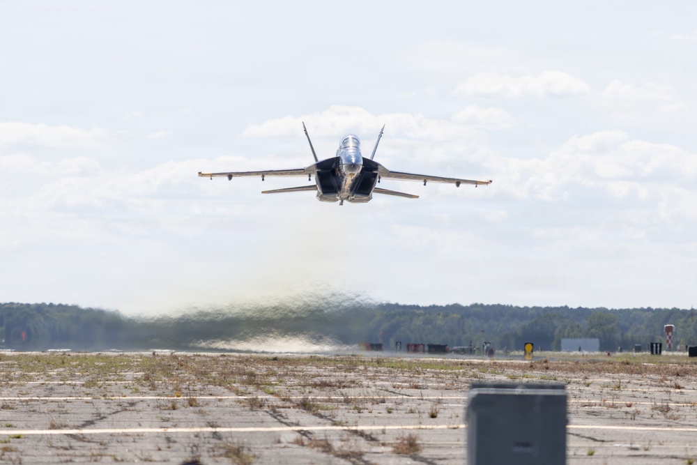 A military jet aircraft, consistent with an F/A-18 Hornet or Super Hornet, is captured in a low-altitude, high-speed pass directly over a runway at what appears to be a military airfield. The aircraft is viewed head-on, flying extremely close to the tarmac surface, generating a visible dust and debris wake beneath it. The surrounding environment includes a flat airfield, tree line in the background, and overcast sky.