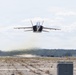 A military jet aircraft, consistent with an F/A-18 Hornet or Super Hornet, is captured in a low-altitude, high-speed pass directly over a runway at what appears to be a military airfield. The aircraft is viewed head-on, flying extremely close to the tarmac surface, generating a visible dust and debris wake beneath it. The surrounding environment includes a flat airfield, tree line in the background, and overcast sky.