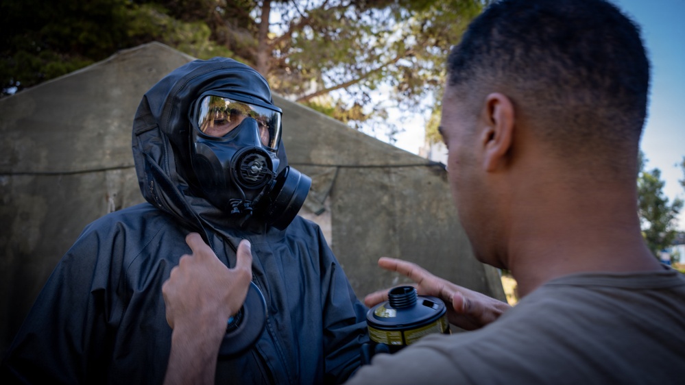 Two individuals are engaged in what appears to be a military or emergency response training exercise involving CBRN (Chemical, Biological, Radiological, Nuclear) protective equipment. One person is fully equipped with a black gas mask and protective suit, while the other in a tan/olive shirt holds a gas mask filter canister and appears to be providing instruction or assistance. The setting is an outdoor area with a concrete structure and trees visible in the background.