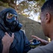 Two individuals are engaged in what appears to be a military or emergency response training exercise involving CBRN (Chemical, Biological, Radiological, Nuclear) protective equipment. One person is fully equipped with a black gas mask and protective suit, while the other in a tan/olive shirt holds a gas mask filter canister and appears to be providing instruction or assistance. The setting is an outdoor area with a concrete structure and trees visible in the background.