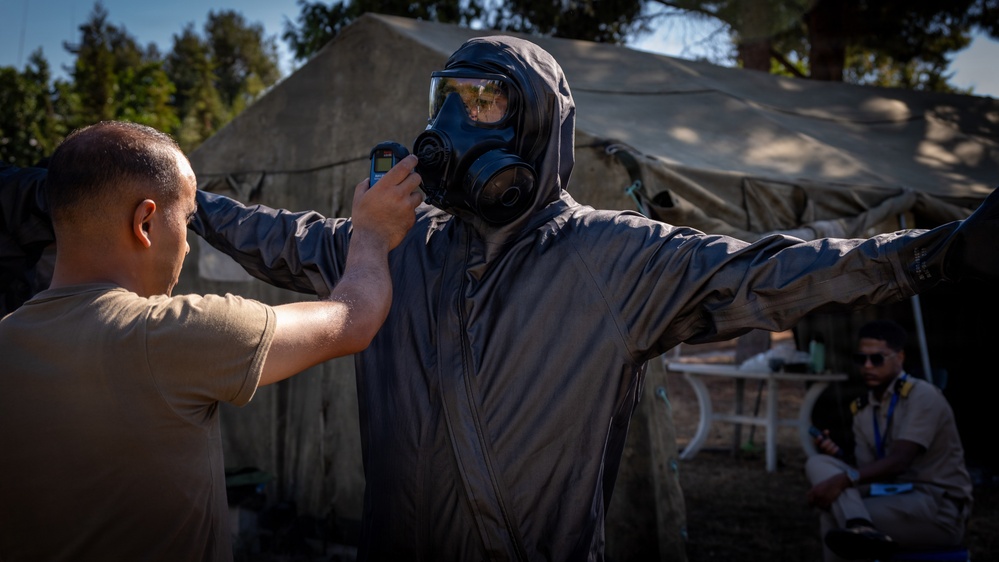 A military or emergency response training exercise is underway outdoors near a field tent. One person in a full CBRN (Chemical, Biological, Radiological, Nuclear) protective suit and gas mask stands with arms outstretched while another individual in a tan uniform scans them with a handheld detection device. A third person in the background appears to be observing or monitoring the exercise.