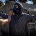 A military or emergency response training exercise is underway outdoors near a field tent. One person in a full CBRN (Chemical, Biological, Radiological, Nuclear) protective suit and gas mask stands with arms outstretched while another individual in a tan uniform scans them with a handheld detection device. A third person in the background appears to be observing or monitoring the exercise.