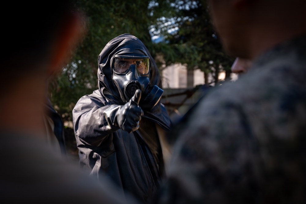 A person wearing full CBRN (Chemical, Biological, Radiological, Nuclear) protective gear, including a gas mask, hood, and rubber gloves, appears to be instructing or briefing a group of individuals in military camouflage uniforms. The central figure is gesturing with a gloved hand toward the audience, suggesting a training or demonstration context. The setting appears to be an outdoor environment with trees visible in the background.
