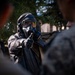 A person wearing full CBRN (Chemical, Biological, Radiological, Nuclear) protective gear, including a gas mask, hood, and rubber gloves, appears to be instructing or briefing a group of individuals in military camouflage uniforms. The central figure is gesturing with a gloved hand toward the audience, suggesting a training or demonstration context. The setting appears to be an outdoor environment with trees visible in the background.